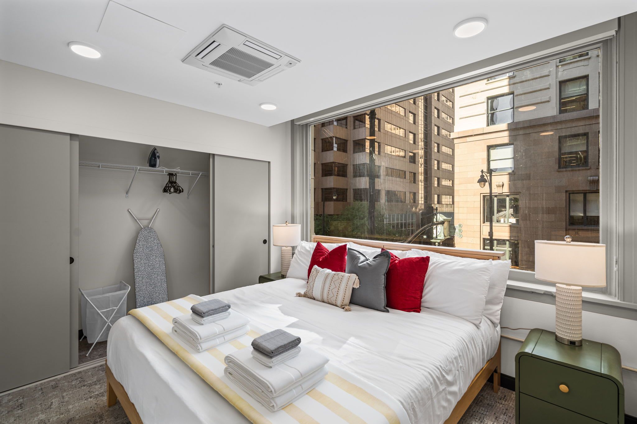 A modern hotel room with a large bed, a wardrobe, a window with a city view, and a small bedside table.