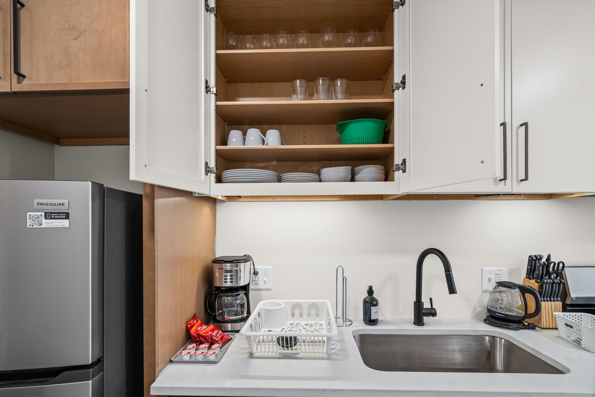A kitchenette with a refrigerator, coffee maker, sink, and cabinets containing dishes and glasses.