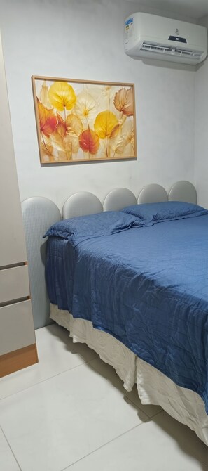 Quarto com cama, ar-condicionado fixado na parede e uma pintura floral emoldurada.