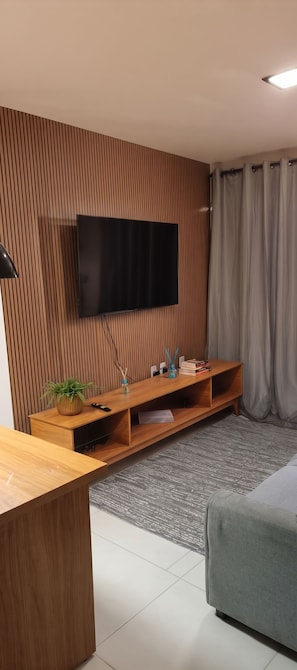 Uma sala de estar moderna com uma TV de tela plana fixada em uma parede de madeira, um rack de madeira, um sofá cinza e uma mesinha com um abajur.