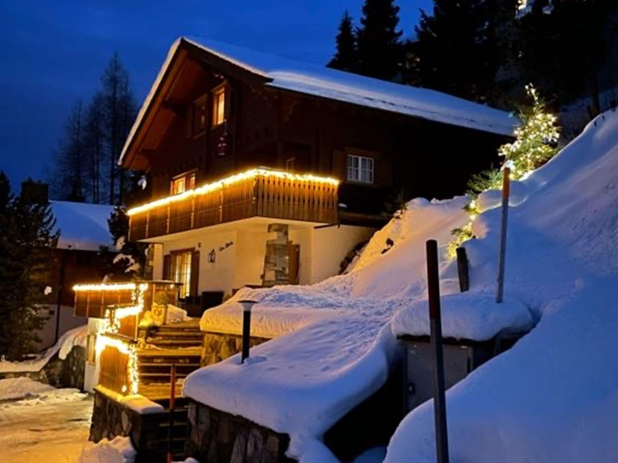 Una cabaña de madera con balcón, escalones cubiertos de nieve y luces colgantes.