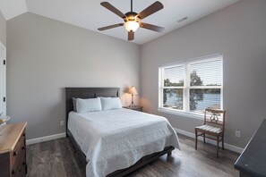 Un dormitorio con cama, mesita de noche, lámpara, silla, ventana con persianas y ventilador de techo.
