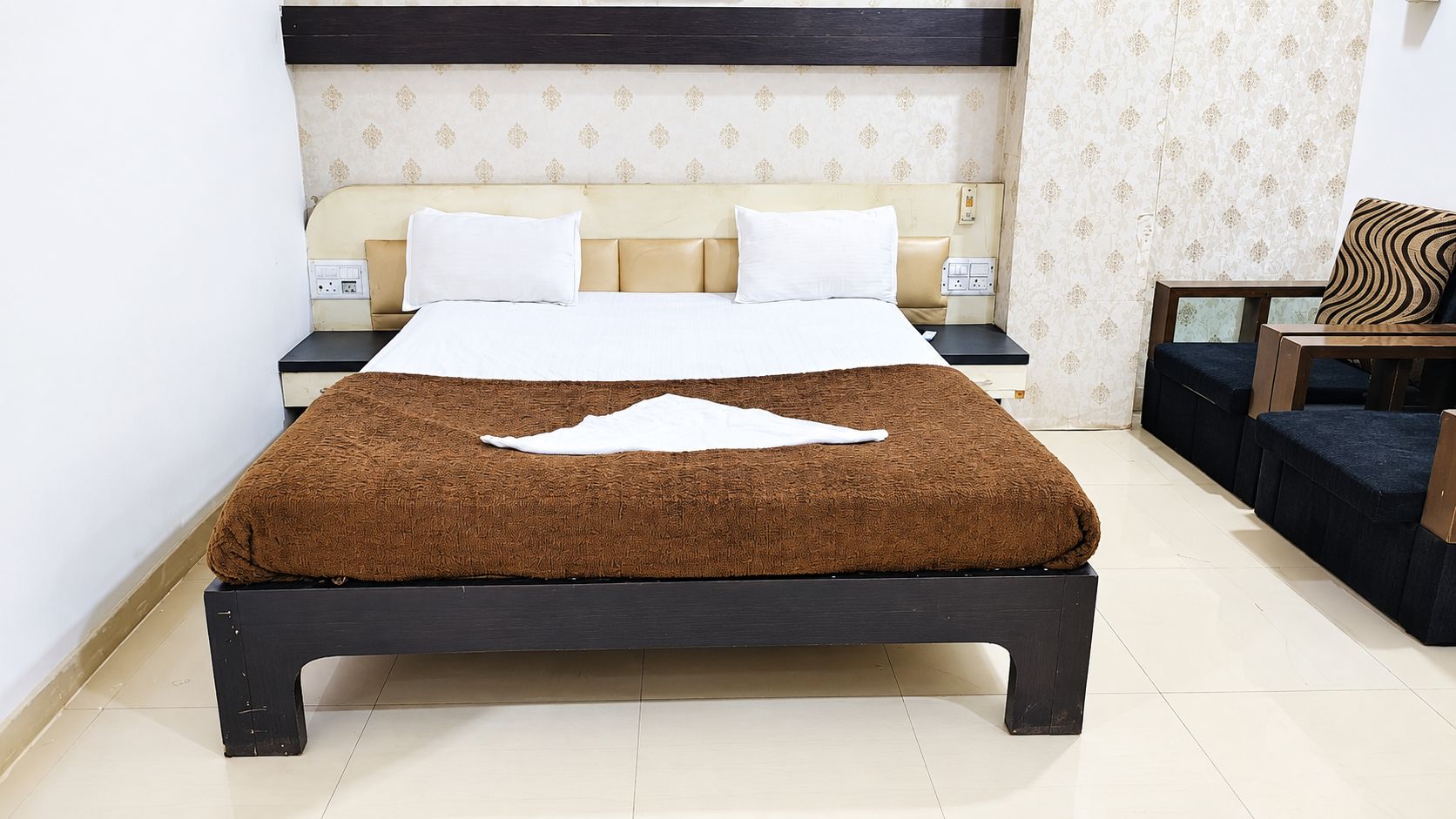 A neatly made bed with a brown blanket and white pillows, a bedside table with a lamp, and a patterned wall.