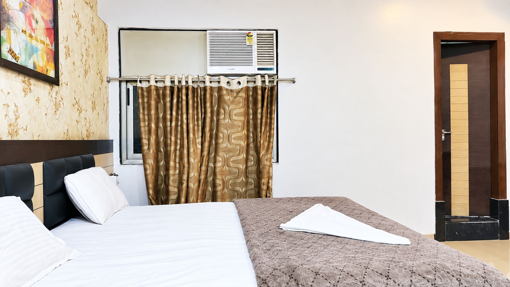 A hotel room with a bed, a window with curtains, an air conditioning unit, and a door.