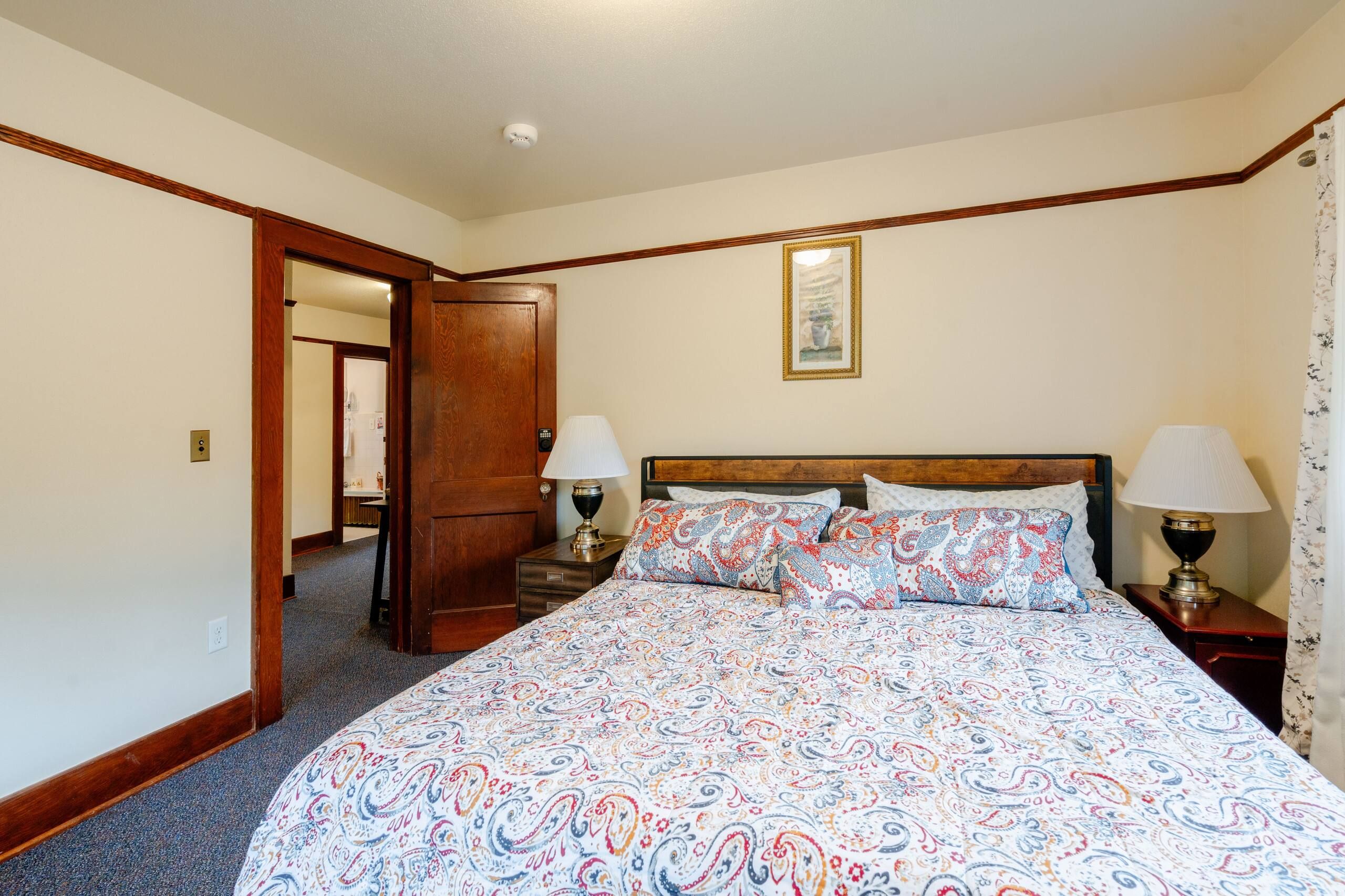 A hotel room with a large bed, two bedside lamps, a door leading to another room, and a framed picture on the wall.