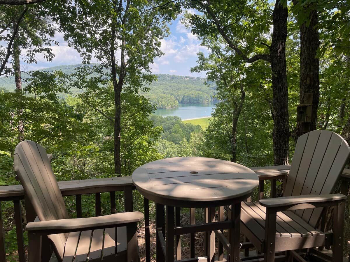 A deck with Adirondack chairs and a table overlooking a lake surrounded by trees.