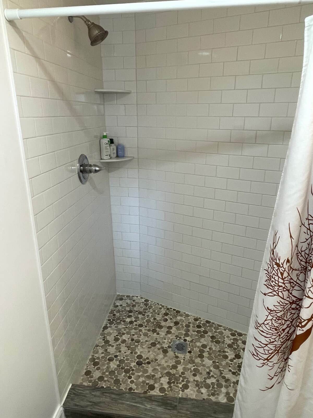 Una doccia con pareti piastrellate di bianco, un tendaggio con motivo alberi e uno scaffale con articoli da toilette.