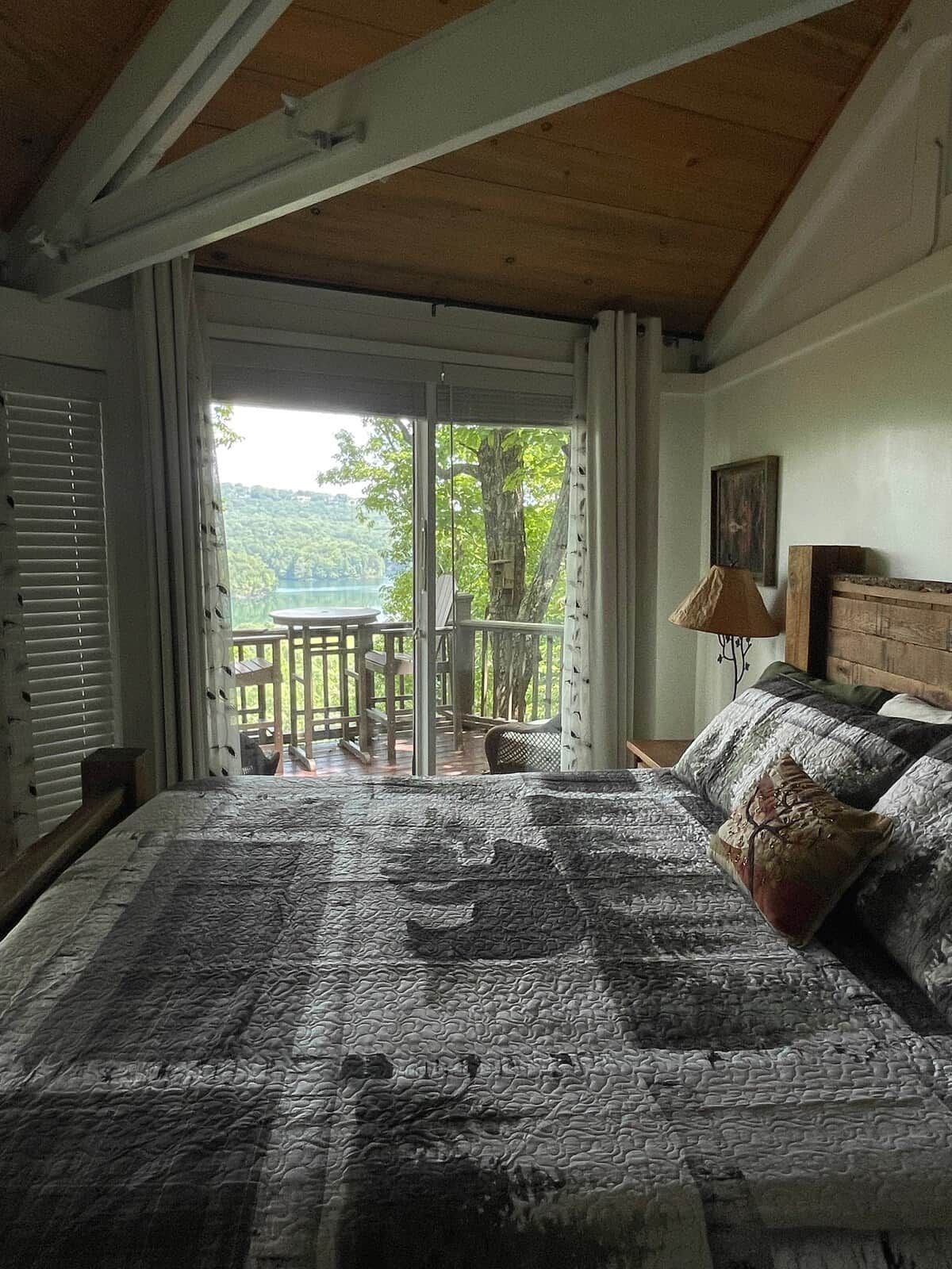 A bedroom with a large bed, a wooden headboard, a nightstand, a lamp, and a view of a balcony with a table and chairs.