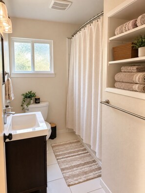 A bathroom with a white sink, a toilet, a shower curtain, a towel rack, and a shelf with folded towels. - Peaceful 4-bedroom house in South Lake Tahoe perfect for families (South Lake Tahoe)