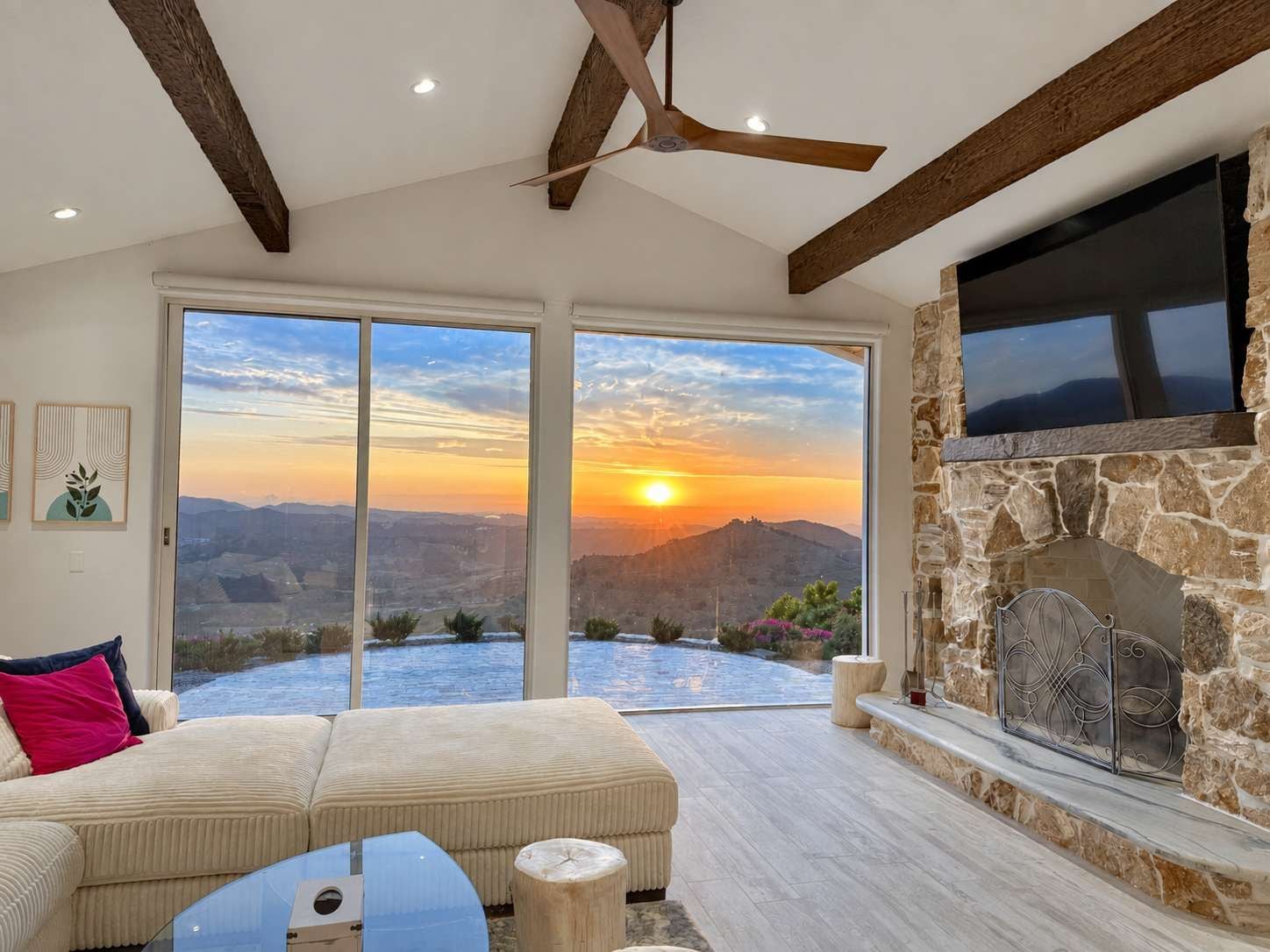 A modern living room with a sofa, a coffee table, and a fireplace. There is a large window offering a view of the sunset and mountains.