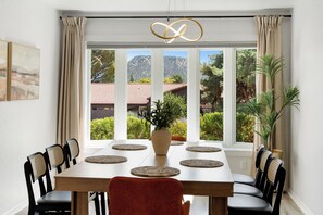 Un coin repas avec une table dressée pour quatre personnes, entourée de chaises, un vase avec une plante et une vue sur une montagne et des arbres, visible à travers de grandes fenêtres.
