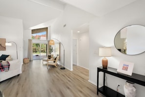 Une chambre d’hôtel moderne avec un plancher en bois, un canapé, un fauteuil, un bureau avec une lampe, un miroir rond et une vue sur l’extérieur.