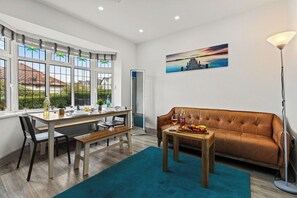 A modern living room with a brown leather sofa, a dining table with chairs, and a large window with a view of a house and greenery. - 4 Doubles, 4 En-Suites & a Big Garden – Sleeps 8 (Ashton-on-Ribble)