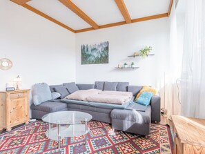Ein modernes Wohnzimmer mit einem grauen Sofa, einem Glastisch, einem gemusterten Teppich, Holzbalken an der Decke und einem Bild an der Wand.
