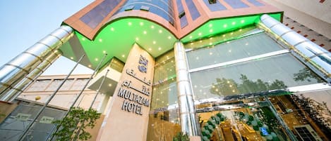 A modern hotel entrance with a prominent sign, large glass windows, and a green-lit facade.
