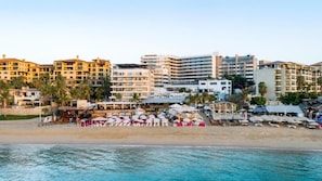 Un hotel a primera línia de platja amb diversos edificis, una platja sorrenca i ombrel·les.