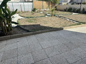 Una zona exterior pavimentada amb un petit jardí, una tanca de formigó i una valla metàl·lica al fons.
