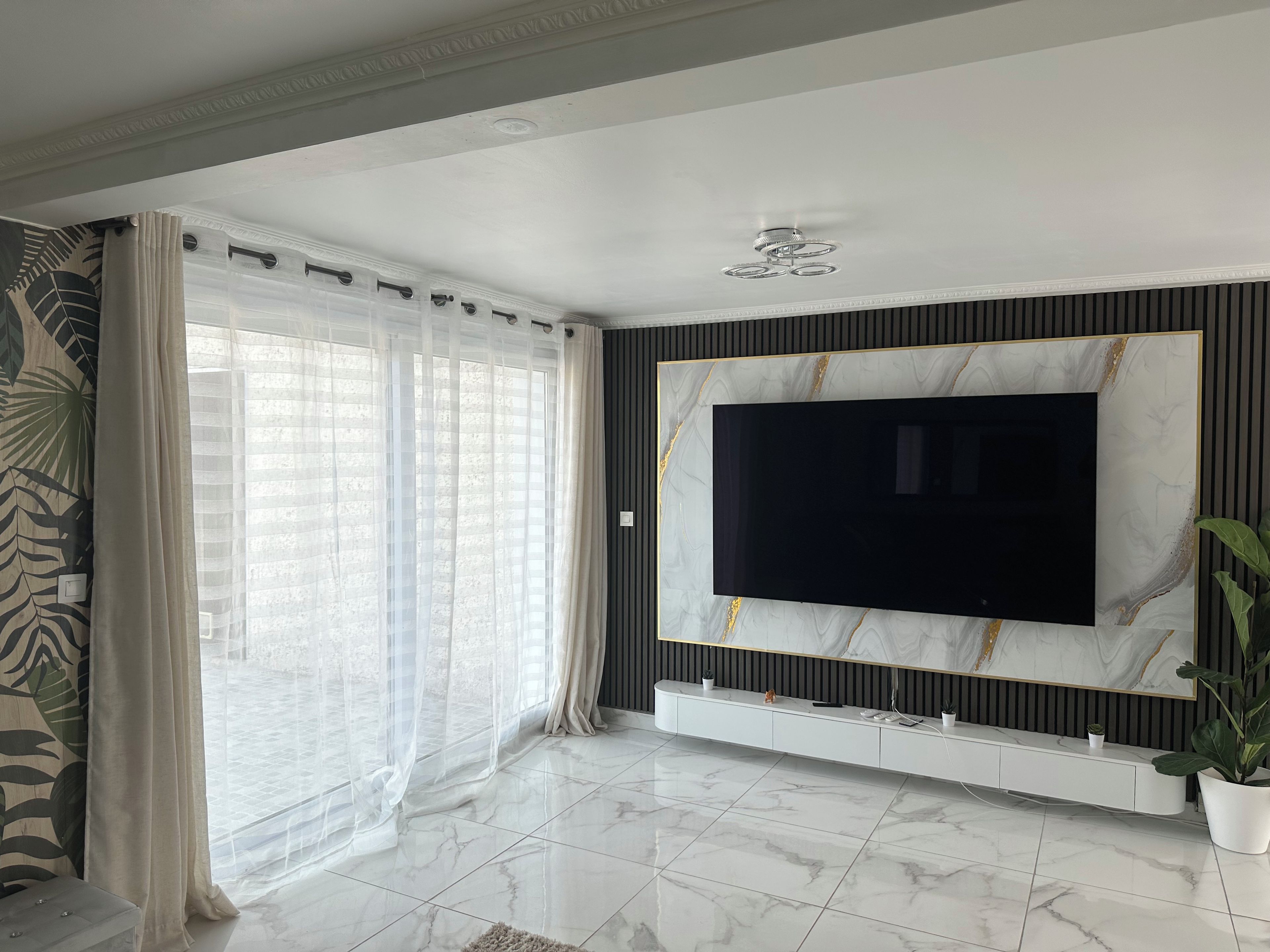 A modern living room with a large flat-screen TV mounted on a marble-patterned wall, a sliding glass door with sheer curtains, and a potted plant.