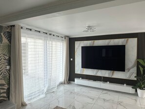 Ein modernes Wohnzimmer mit einem großen Flachbildfernseher an einer Wand mit Marmoroptik, einer Glastür mit durchsichtigen Vorhängen und einem Topf mit Pflanze.