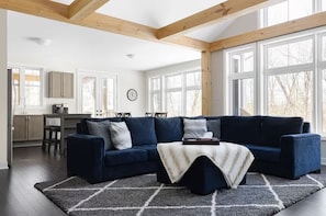 Un salon moderne avec un canapé d’angle bleu foncé, une table basse et un tapis. On aperçoit à l’arrière-plan un coin cuisine avec des placards et une table à manger.