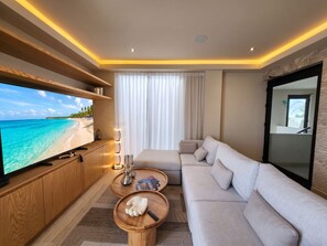 A modern living room with a large flat-screen TV, a sofa, a coffee table, and a view of a beach. - Ameth- Apartments w/ Rooftop Pool & Gym (Cabo San Lucas)