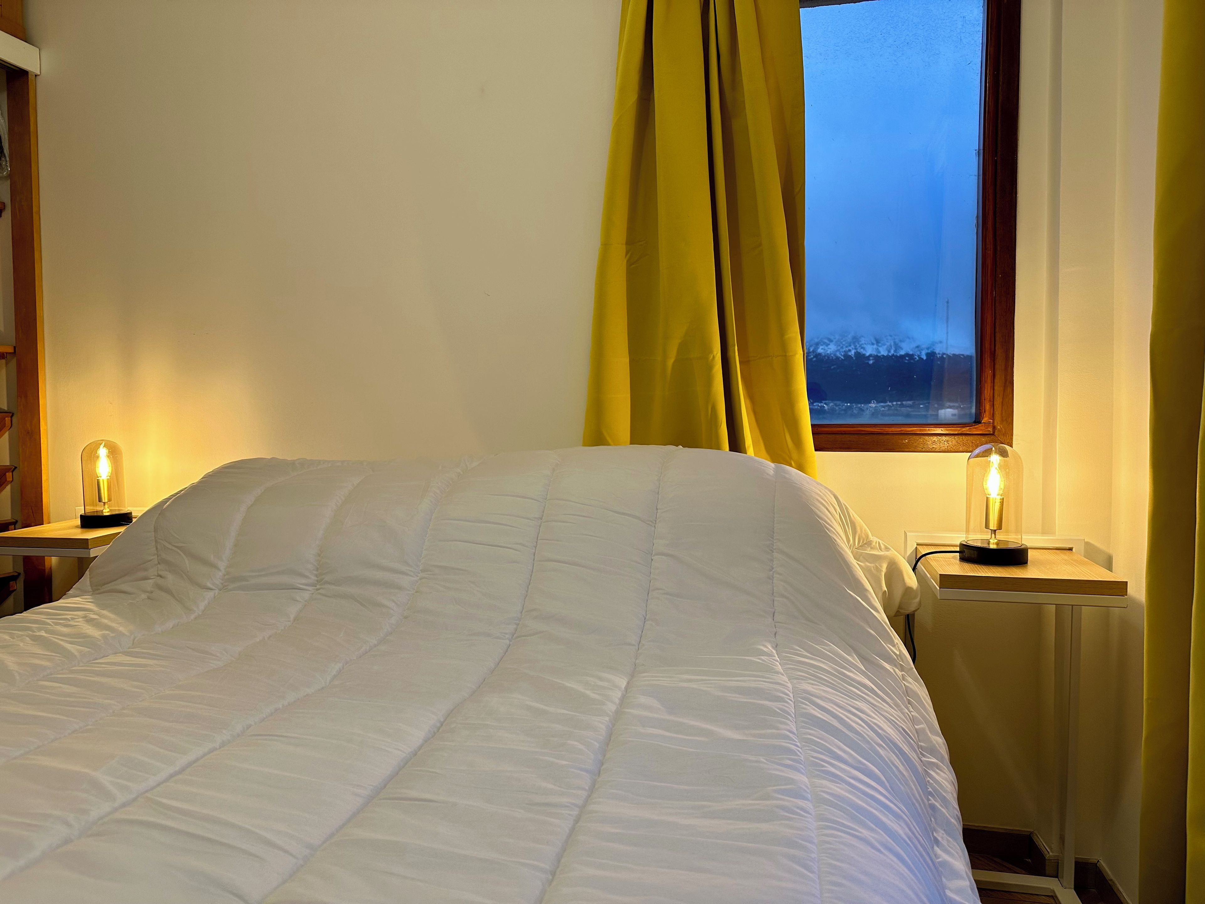 Quarto com cama, duas luminárias de cabeceira, janela com cortinas e vista para o exterior.