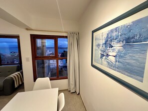 A room with a large window, a framed painting of boats, a sofa, and a dining table. - Shackleton Apartamento (Ushuaia)