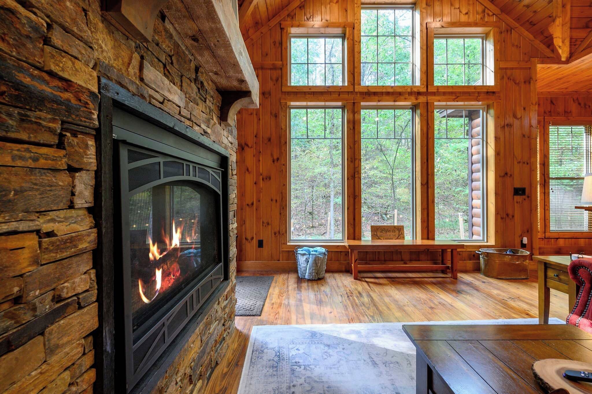 Ein Interieur einer Blockhütte mit Kamin, Holzböden und großen Fenstern mit Blick auf den Wald.