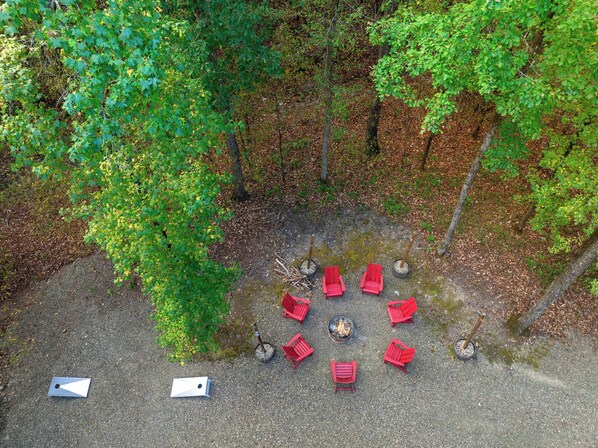 Vista aérea de un camping con sillas rojas dispuestas alrededor de un brasero central, rodeado de árboles y un sendero de grava.