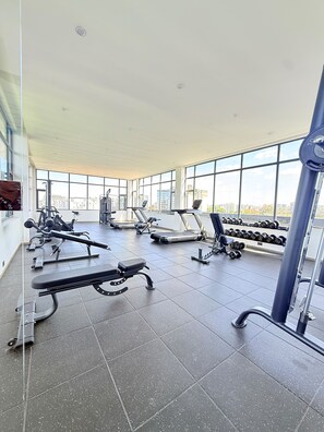 Un gimnasio con diverso equipo de ejercicio, ventanas amplias y suelo de baldosas.