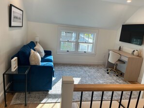 Une pièce avec un canapé bleu, un bureau avec une chaise, un tableau encadré au mur et une fenêtre donnant sur un paysage.