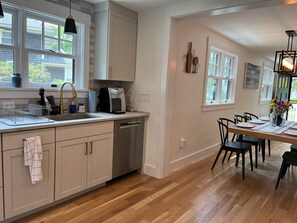 A modern kitchen with a dishwasher, a coffee maker, and a dining area with a table set for two. - A quiet Oak Bluffs retreat steps from Circuit Avenue and Inkwell Beach. (Oak Bluffs)