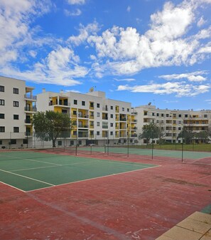 An outdoor tennis court with a red and green surface, surrounded by a fence, and adjacent to a multi-story residential building with balconies and satellite dishes. - 1-Bedroom Apartment (Armação de Pêra)