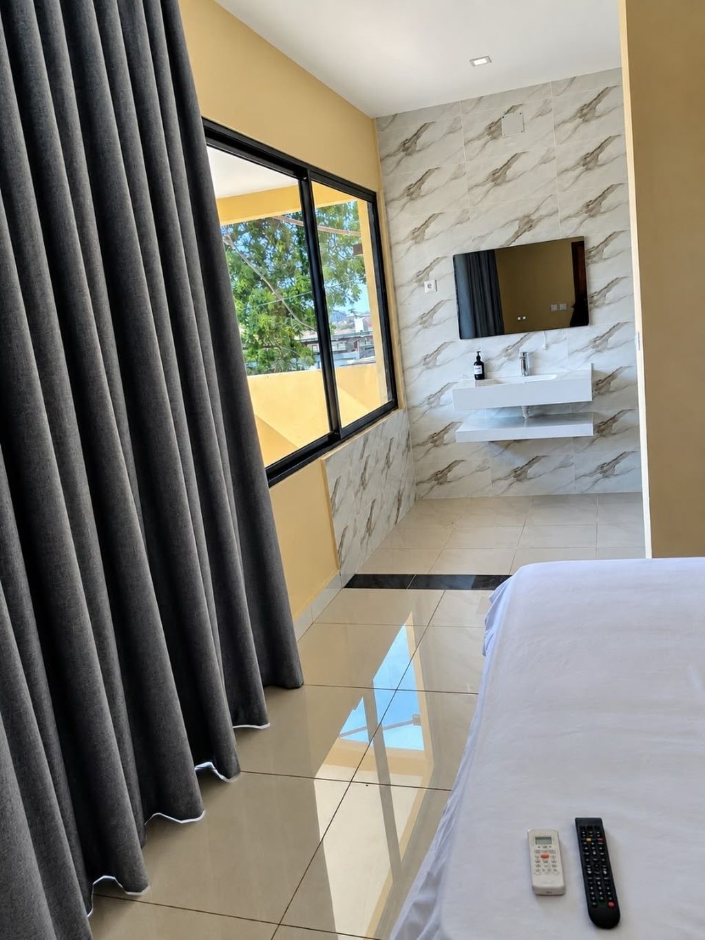 A hotel room with a bed, a TV mounted on the wall, a bathroom with a mirror, and a window with curtains.