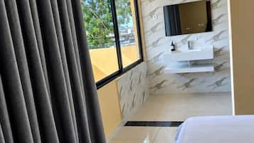 A hotel room with a bed, a TV mounted on the wall, a bathroom with a mirror, and a window with curtains.