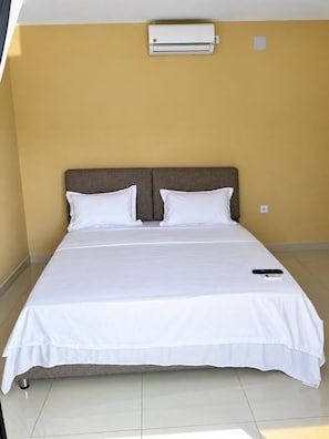 Un llit individual amb matalàs i almoadines blancs, un comandament a distància i un aparell d'aire condicionat fixat a la paret en una habitació amb parets grogues i terra de rajola.