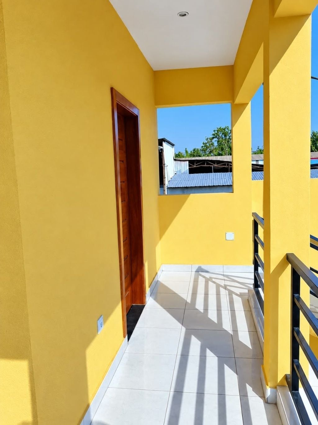 A corridor with yellow walls, a wooden door, and a view of a building with a corrugated metal roof.