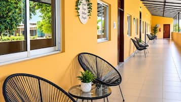 A corridor with black chairs and a potted plant, a wall with a sign, and windows with views of greenery.