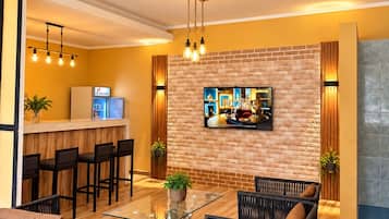 Un salon moderne avec une table basse en verre, des meubles en osier, un téléviseur à écran plat fixé à un mur en briques et un coin cuisine avec des tabourets de bar.
