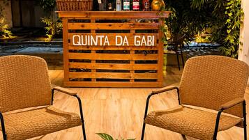 Zona de recepción con mostrador de madera que dice 'QUINTA DA GABI', dos sillas de mimbre, una mesa redonda con una planta y un fondo de follaje e iluminación colgante.