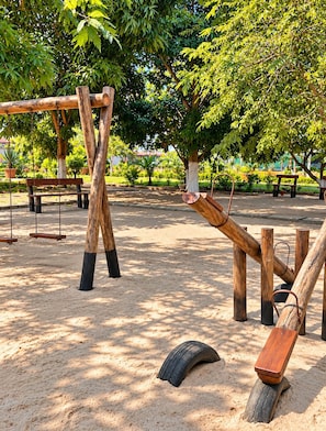 Un parc infantil amb cadira d'oscalls i neumàtics penjants, envoltat d'arbres i bancs.