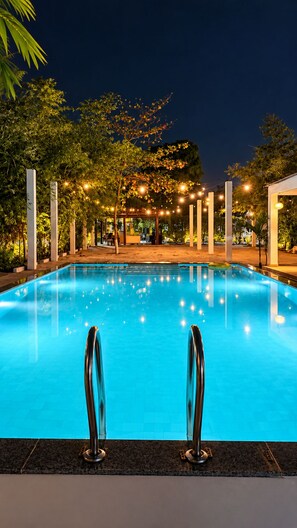 Una piscina con agua azul cristalina, rodeada de árboles y columnas, con una zona de estar cubierta al fondo.
