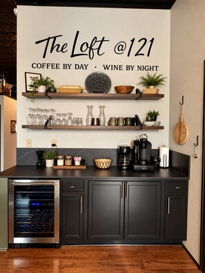 Eine Küche mit einem Schild, auf dem 'The Loft @121 COFFEE BY DAY WINE BY NIGHT' steht, Regalen mit Glaswaren, einer Kaffeemaschine und einem Weinkühler.