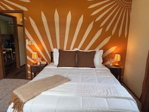 Una cama bien hecha con edredón blanco y dos almohadas marrones, flanqueada por dos mesitas de noche con lámparas, en una habitación con diseño de sol radiante en la pared.