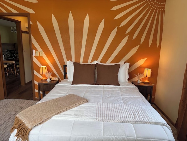 Un lit bien fait, recouvert d’une couette blanche et agrémenté de deux oreillers marron, flanqué de deux tables de chevet avec des lampes, dans une pièce dont le mur est décoré d’un motif en forme de soleil.