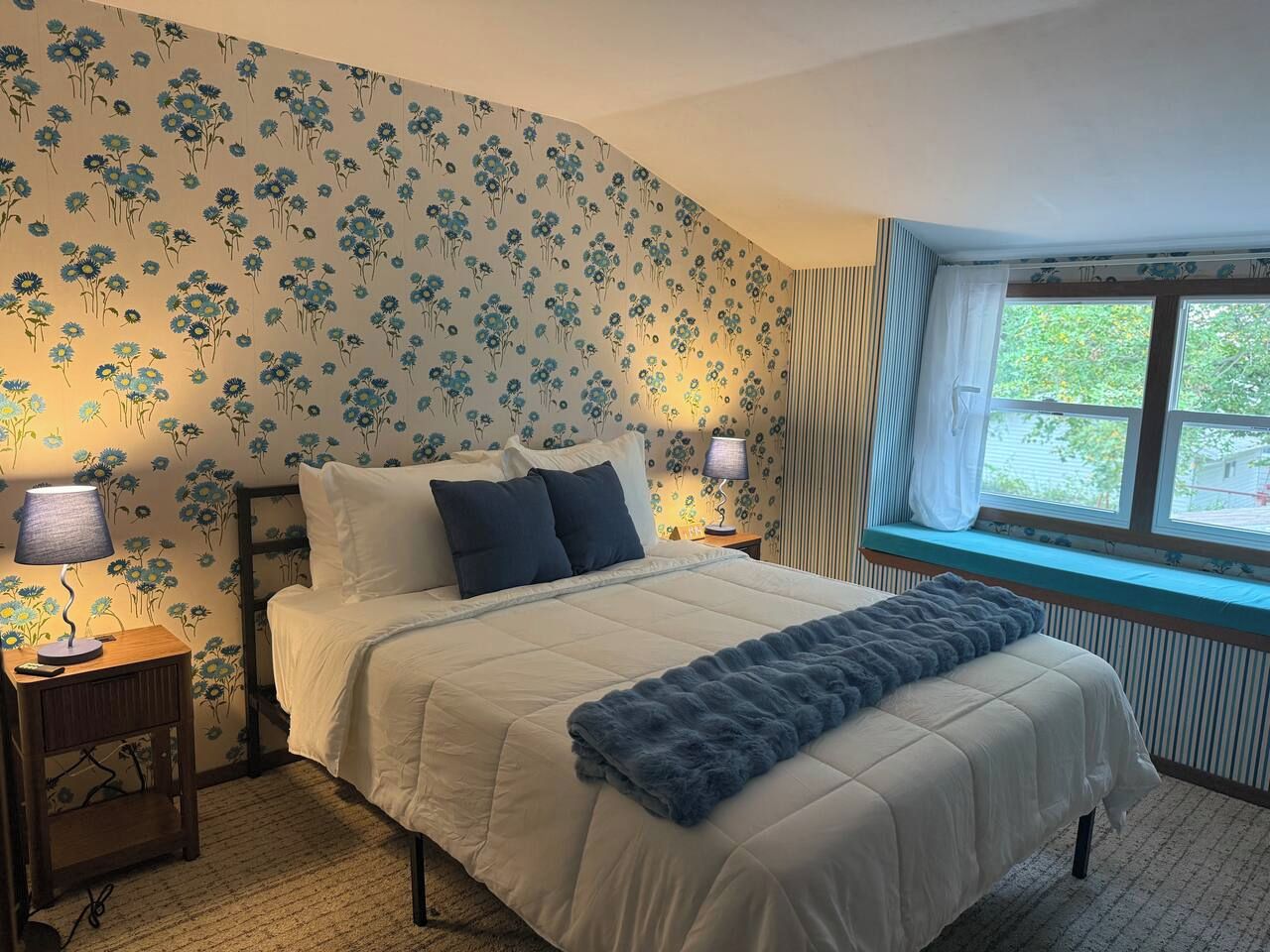 Un dormitorio con cama, dos mesitas de noche, una ventana con vistas y papel tapiz floral.