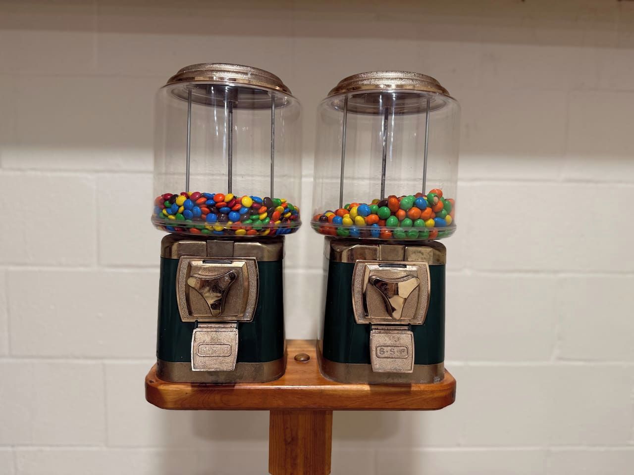 Two vintage candy dispensers with transparent tops and metallic bases, filled with colorful candies, mounted on a wooden stand against a white wall.