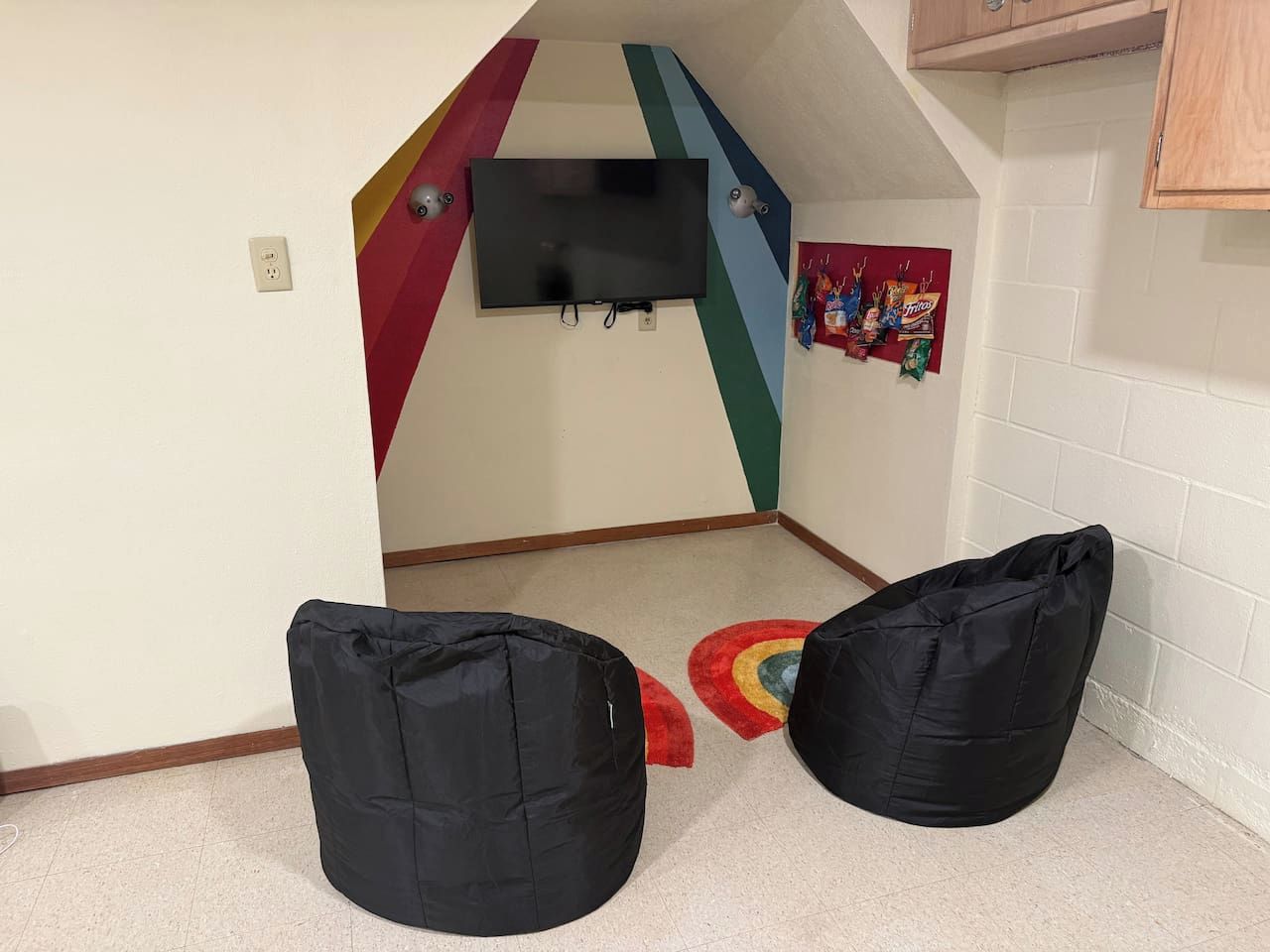 Quarto com televisão na parede, dois puffs pretos e um tapete colorido.