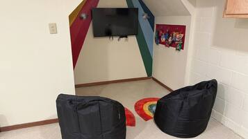 Quarto com televisão na parede, dois puffs pretos e um tapete colorido.