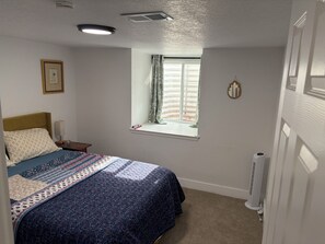Ein Schlafzimmer mit einem Bett, einem Fenster mit Vorhängen, einem Nachttisch und einer Wandleuchte.